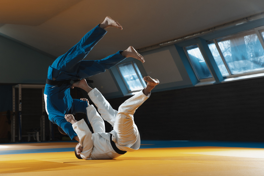 Dwóch zawodników judo podczas dynamicznej akcji na macie. Jeden z nich w białym kimonie wykonuje rzut, unosząc drugiego zawodnika w niebieskim kimonie w powietrze. Sala treningowa z dużymi oknami w tle zapewnia odpowiednie oświetlenie do treningu. Scena ukazuje intensywność i precyzję techniki judo.