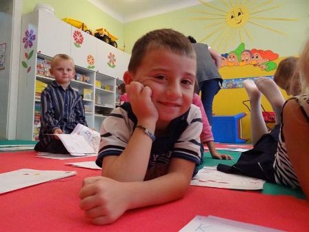 Chłopiec leży na podłodze w przedszkolu, uśmiechając się do kamery. Przed nim leżą kolorowanki i rysunki. W tle widoczne są inne dzieci i dekoracje ścienne z motywami słonecznymi. Scena przedstawia dzieci zaangażowane w kreatywne zajęcia w radosnym i kolorowym środowisku przedszkolnym.